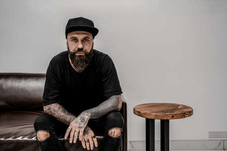Young male tattoo artist with beard looks serious sitting on couch in workshop place.の写真素材