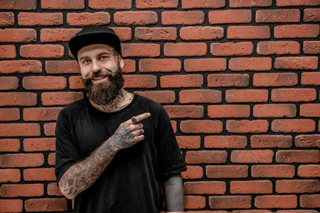 Handsome old-fashioned hipster in t-shirt and cap, pose and smile. On a brick background.の写真素材