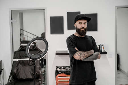 Young male tattoo artist with beard looking positive and happy standing and smiling in workshop place.の写真素材