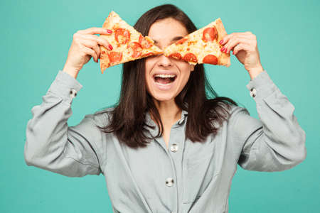 Photo of cute lady covers eyes up with pizza. Wears grey shirt, isolated turquoise color backgroundの写真素材