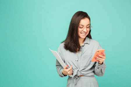 Photo of cute business lady in glasses holds papers and texting on phone. Wears grey shirt, isolated turquoise color backgroundの写真素材