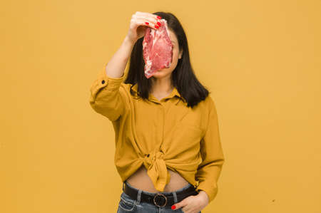 Concept photo of sweet female covered her face up bu meat. Wears yellow shirt, isolated yellow color backgroundの写真素材
