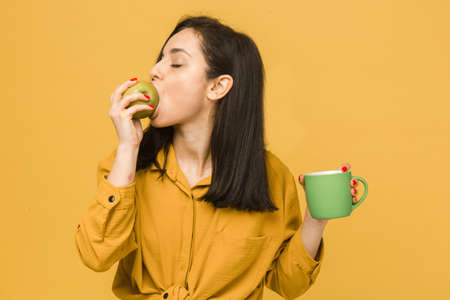 Concept photo of young female eating apple and drinking something. Wears yellow shirt, isolated yellow color backgroundの写真素材