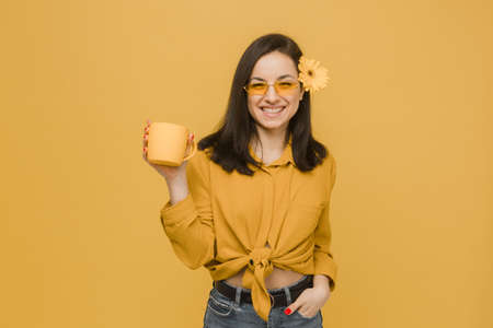 Concept photo of young female in glasses and flower in hair, holds drink, feeling spring time. Wears yellow shirt, isolated yellow color backgroundの写真素材