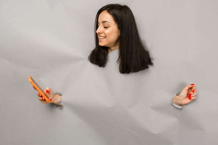 Concept photo of young female tearing paper and peeking out hole, holding smartphone, isolated grey color backgroundの写真素材