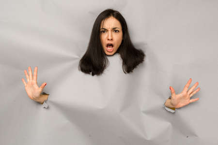 Concept photo of young female tearing paper and peeking out hole, shocked about commercial offer on copy space, isolated grey color backgroundの写真素材