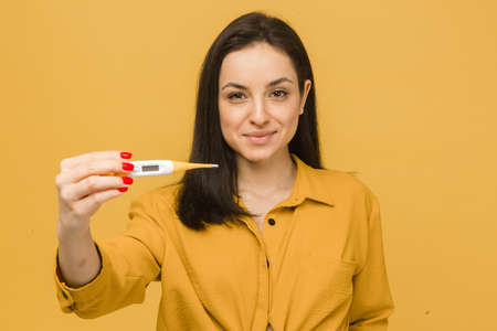 Concept photo of cute female in face mask holds thermometer. Wears yellow shirt, isolated yellow color backgroundの写真素材