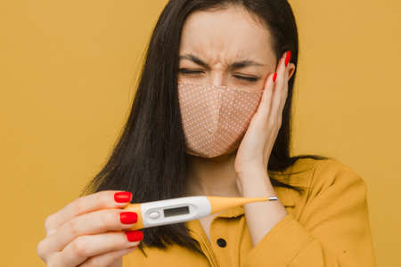 Concept photo of sick female in face mask with headache holds thermometer. Wears yellow shirt, isolated yellow color backgroundの写真素材