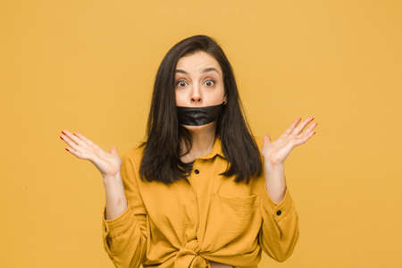 Concept photo of female victim with her mouth taped up, holds her head. Wears yellow shirt, isolated yellow color backgroundの写真素材