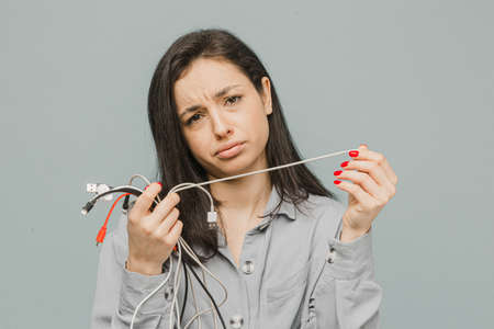 Photo of cute female disappointed of broken chargers. Wears grey dress, isolated grey color backgroundの写真素材