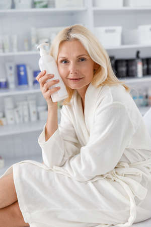 Photo senior Caucasian female in white bathrobe, holding lotion jar in beautician salonの写真素材