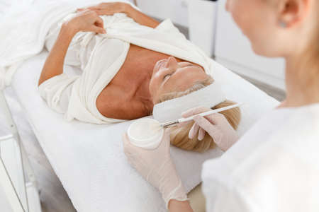 Close up face peeling mask, spa beauty treatment, skincare. Senior woman getting facial care by beautician at spa salon, side viewの写真素材