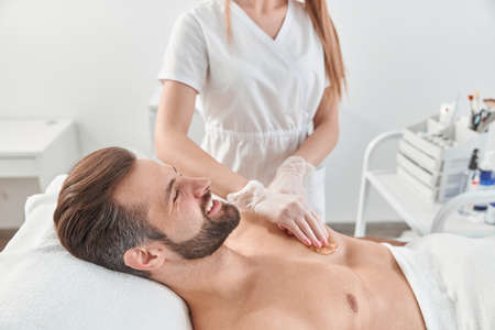 Woman beautician put depilatory wax to chest of young man for hair removal. Depilation with waxの写真素材