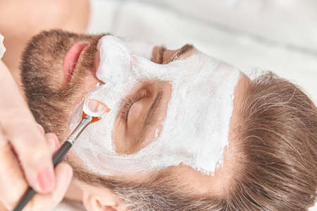Close up bearded young man is relaxing while cosmetologist is spreading white clay on his faceの写真素材