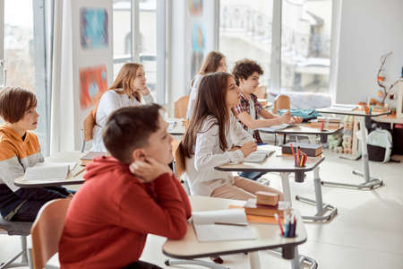 Nice pupils are listening carefully to their tutor. Elementary school kids sitting on desks and reading books in classroomの写真素材
