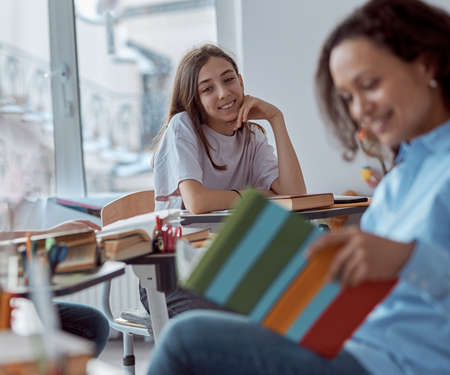 Close up cute schoolgirl smiling while young tutor is reading her. Elementary school kids sitting on desks in classroomの写真素材