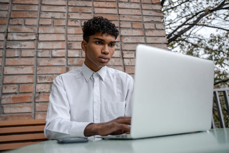 Cheerful man works at a laptop. A black guy sits on a summer terrace or veranda near a brick wall....