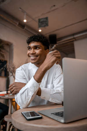 A young black man is sitting at the table with a laptop, drinking tea and using a smartphone. The guy in the cafe. Work outside the office, remote work or study concept. Vertical photo.の写真素材
