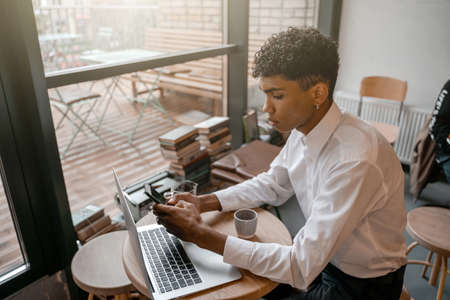 A young black man is sitting at the table with a laptop, drinking tea and using a smartphone. The guy in the cafe. Work outside the office, remote work or study concept.の写真素材
