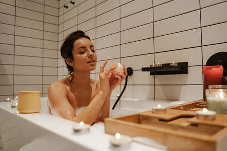 Young woman using mask on face, relaxing in the cosy bath full of foam and decorated with candles. Healthy treatment and enjoyment.の写真素材