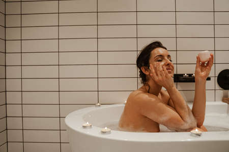 Young woman using cream on face, relaxing in the cosy bath full of foam and decorated with candles. Healthy treatment and enjoymentの写真素材