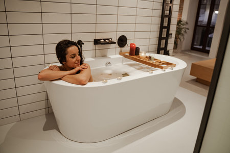 Young and calm woman relaxing in the cosy bath full of foam decorated with candles. Healthy...