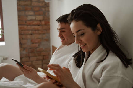 Morning of happy young couple in white bathrobes in bed at home, using phonesの写真素材