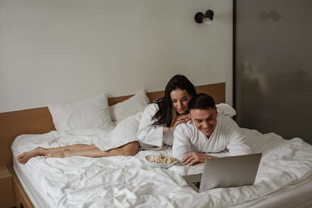 Happy young couple in white bathrobes at home in bed with pop corn, watching movie on laptop and laughingの写真素材