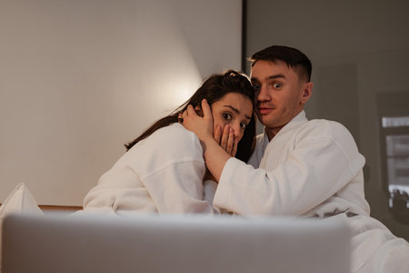 Happy young couple in bathrobes at home with watching horror movie on laptop, woman is scared
