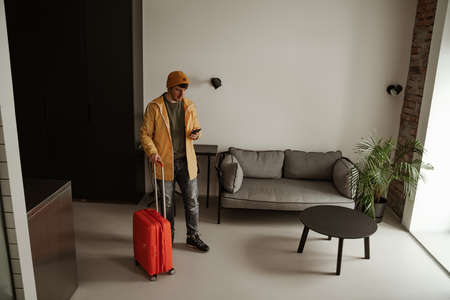 Young man traveler with luggage and phone in hand, standing in hotel room on summer vacation. Cool male in casual clothing with luggageの写真素材