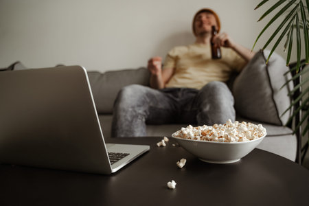 Close up, young man tourist in yellow hat drinking beer with pop corn, watching movie on laptop in hotel room. Male on vacation.の写真素材