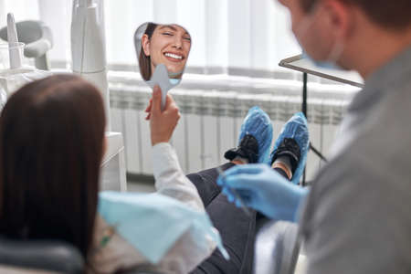 Happy young woman smiling checking out her perfect healthy teeth in the mirror, at the dentist officeの写真素材