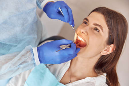 Portrait of young dentist in gloves examining teeth of female client in the hospitalの写真素材