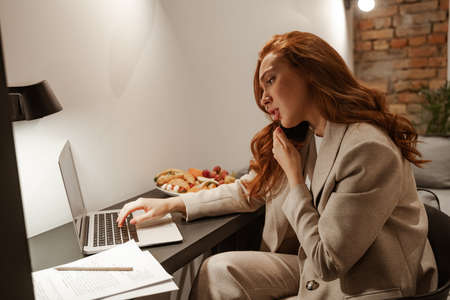 Work at home, business trip. Multitasking concept. A very busy red-haired young woman in a stylish business suit and talking on the phone uses papers and a laptop. She is eating while working.の写真素材