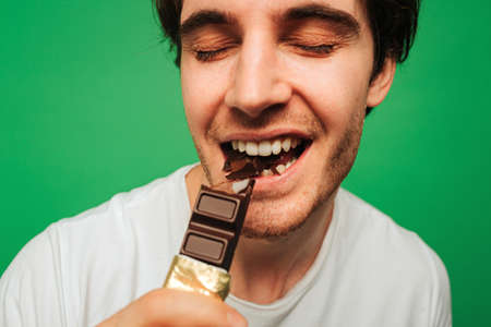 Young man bites chocolate bar and enjoy it, isolated on green backgroundの写真素材