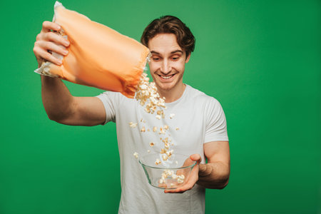 Young man pours popcorn into a plate, isolated on green background