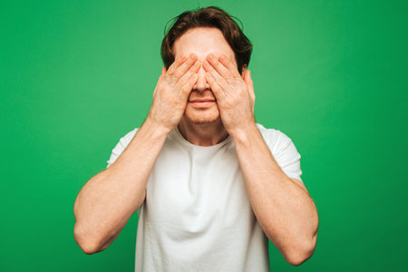 Young man covers eyes up with hands, isolated on green background