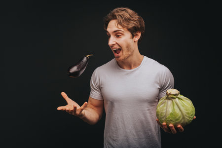 Young blond man in white t-shirt is tossing eggplant up and holding cabbage, isolated over black backgroundの写真素材