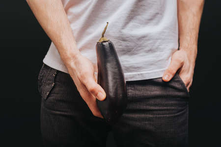 Young blond man in white t-shirt is holding eggplant near the pants, isolated over black backgroundの写真素材