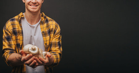 Young blond man in yellow shirt is holding mushrooms in hands with smile, isolated over black...