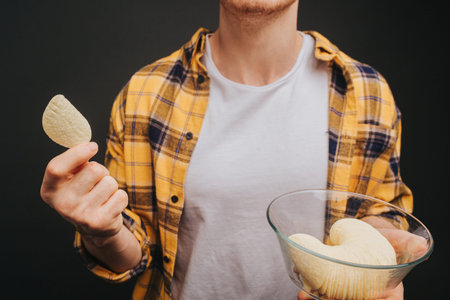 Close up, young blond man in yellow shirt is holding plate with chips, isolated over black...