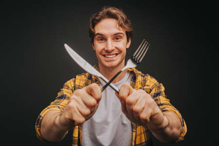 Young blond man in yellow shirt is smiling while holding fork and knife, isolated over black backgroundの写真素材