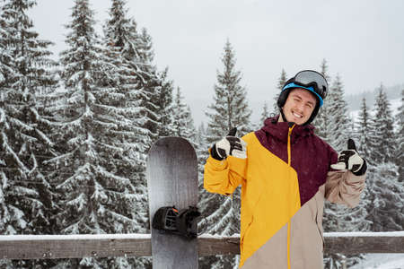 Man in ski equipment, wearing safety glasses shows thumbs up, stands against mountain and trees. Winter sport and recreation, leisure outdoor activitiesの写真素材