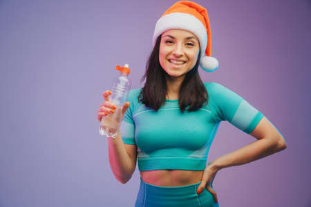 Young sportive woman in Christmas hat training and drinking water, isolated on gradient studio background in neon light. Modern sport, action, motion, youth conceptの写真素材