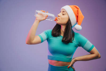 Young sportive woman in Christmas hat training and drinking water, isolated on gradient studio background in neon light. Modern sport, action, motion, youth conceptの写真素材