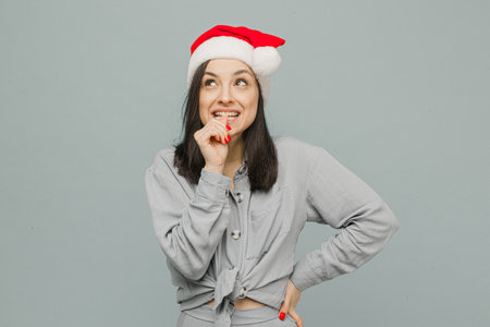 Photo of cute smiling female in Christmas hat looks curious. Wears grey shirt, isolated grey color backgroundの写真素材