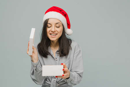 Photo of cute smiling female in Christmas hat is opening present. Wears grey shirt, isolated grey color backgroundの写真素材