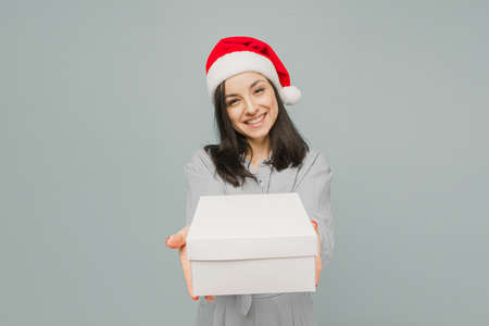 Photo of cute smiling female in Christmas hat giving a present. Wears grey shirt, isolated grey color backgroundの写真素材