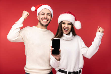 Photo of sweet couple in Christmas hats, holds new smartphone for gift. Male and female are in love, look blessed and smile, isolated over red backgroundの写真素材