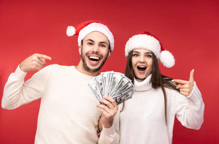 Photo of sweet couple in Christmas hats, happy about holidays, holds money in dollars. Male and female are in love, look blessed and smile, isolated over red backgroundの写真素材